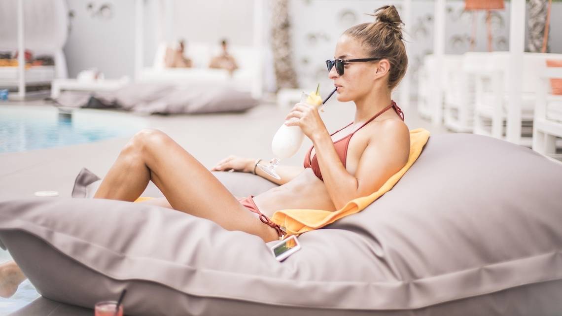 Frau im roten Bikini sitzt mit einem Getränk auf einem grauen Loungesofa am Pool der Ibiza Sun Apartments.