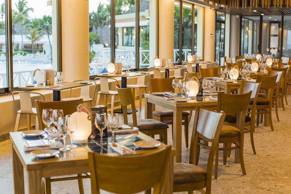 Innenbereich eines Restaurants mit mehreren Tischen und Stühlen, umgeben von großen Fenstern mit Blick auf einen Pool.