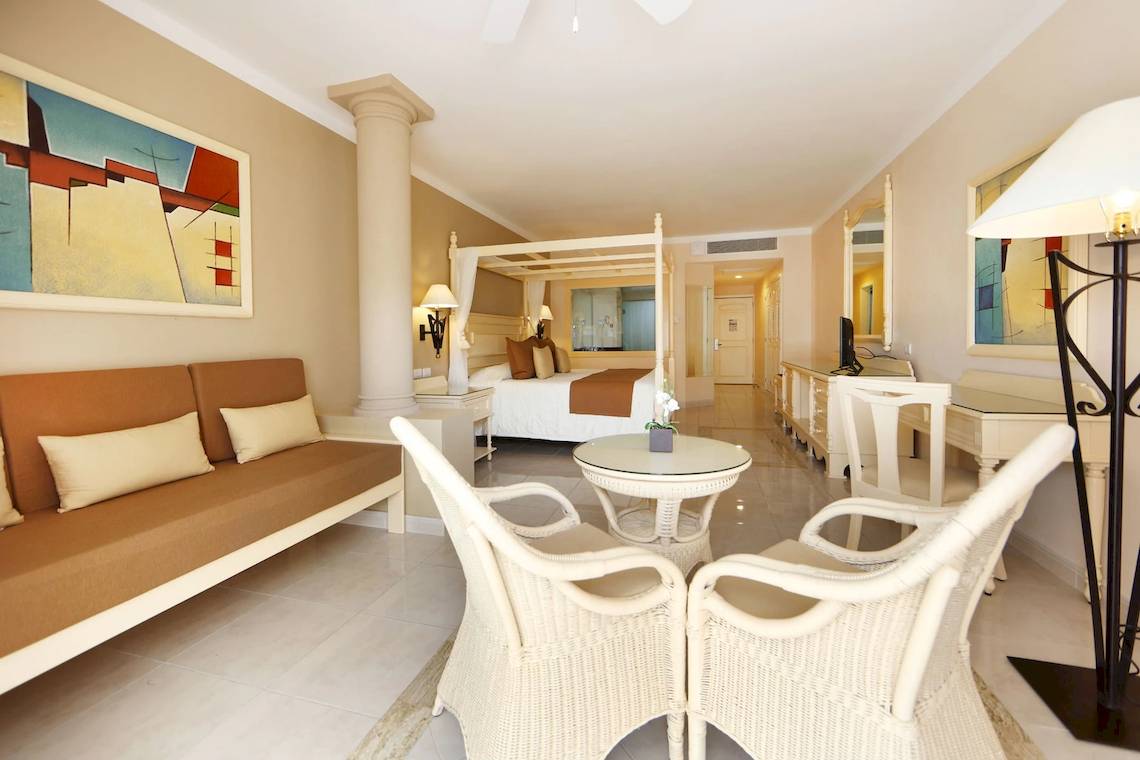Inneneinrichtung einer Hotelsuite im Bahia Principe Luxury Akumal mit einem Bett, Sofa, Tisch und Stühlen.