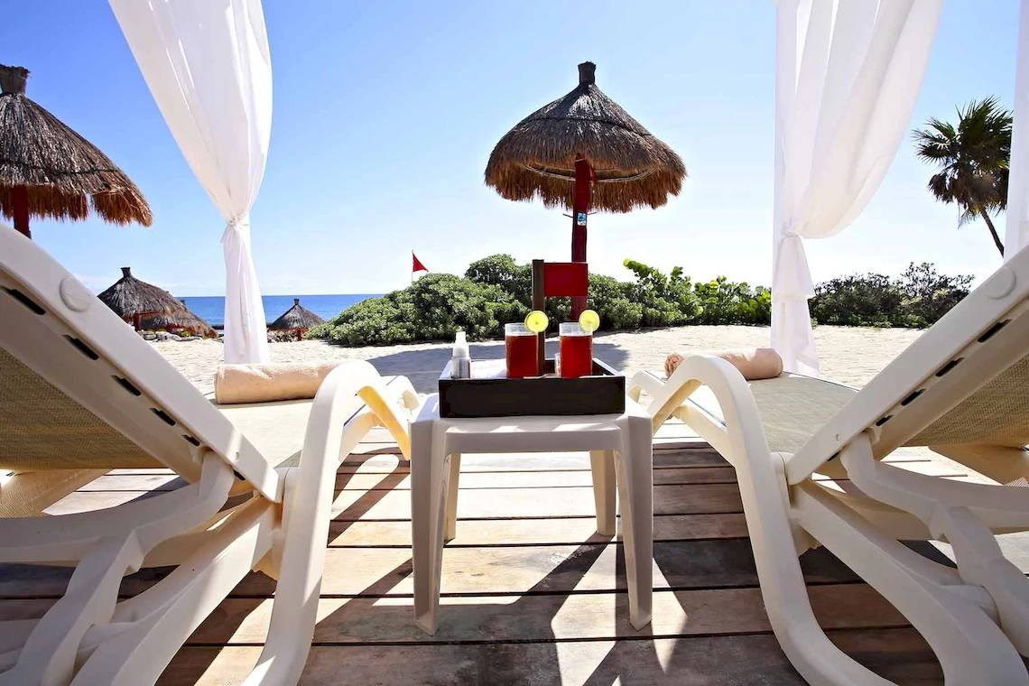 Zwei Sonnenliegen mit Getränken auf einem Tisch auf einem Holzdeck am Strand des Bahia Principe Luxury Akumal