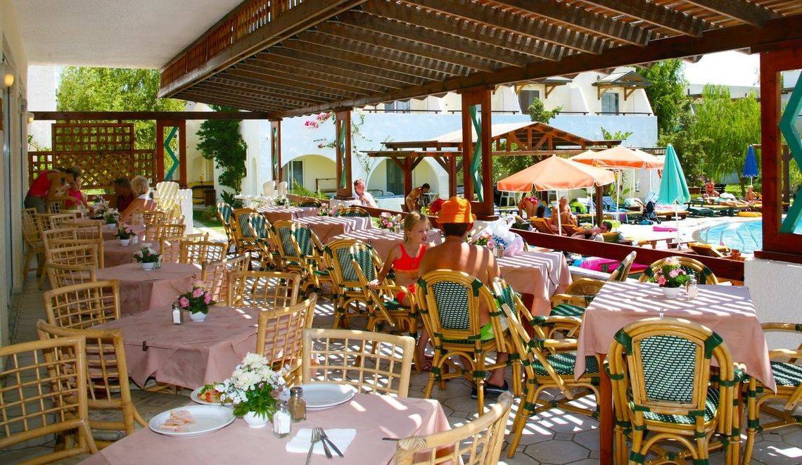 Terrasse des Gaia Village Hotels mit Gästen, Tischen und Sonnenschirmen neben einem Pool.