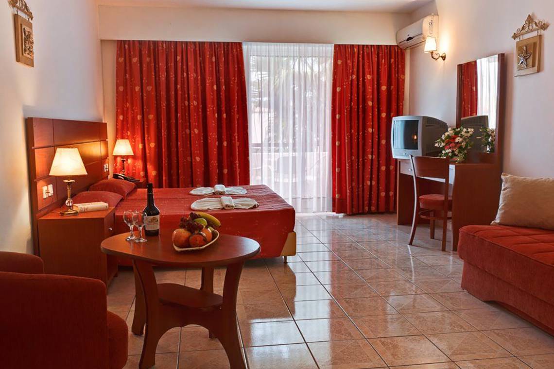 Innenansicht eines Hotelzimmers im Gaia Village Hotel mit einem Doppelbett, einem Tisch mit Obst, einem Fernseher und einem Schreibtisch.