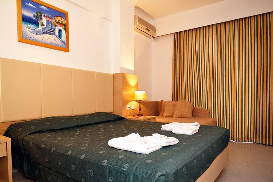 Schlafzimmer im Gaia Village Hotel mit Bett, Kissen, Decken, Nachttisch, Lampe und Klimaanlage.