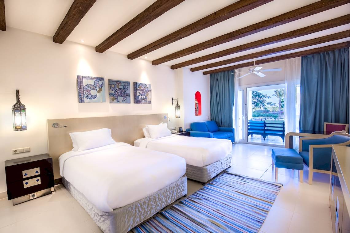 Doppelzimmer im Hilton Marsa Alam Nubian Resort mit zwei Betten, einem Nachttisch, einer Lampe, drei Gemälden an der Wand und einem Balkon.