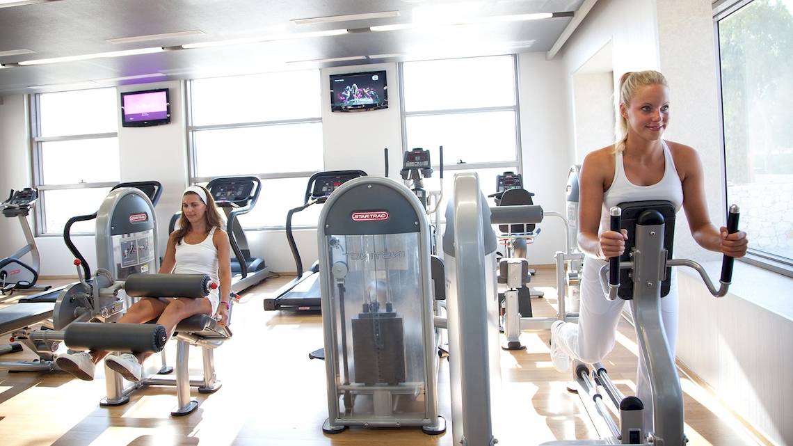 Zwei Frauen trainieren in einem Fitnessstudio, eine auf einem Stepper, die andere auf einem Beinpressegerät.