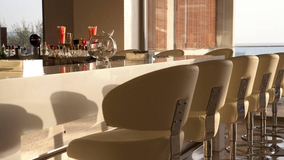 Hotelbar mit Sitzgelegenheiten, Blick auf das Meer und verschiedene Getränkeflaschen auf dem Tresen.