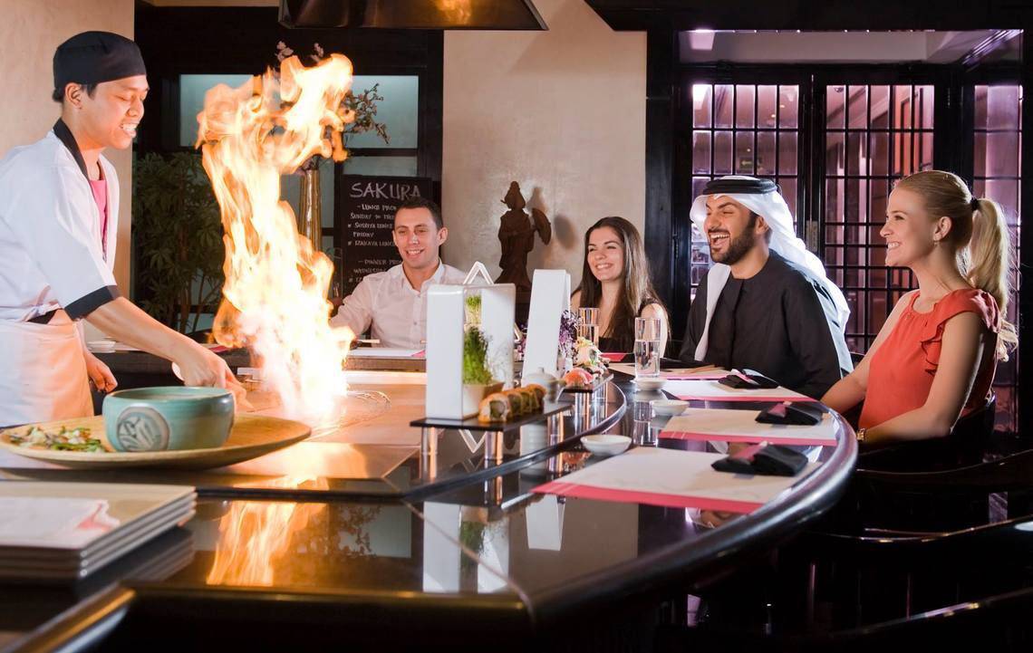 Gäste sitzen an einem Tisch im Millennium Plaza Downtown Sushi Bar und beobachten den Koch, der ein Gericht mit Feuer zubereitet.