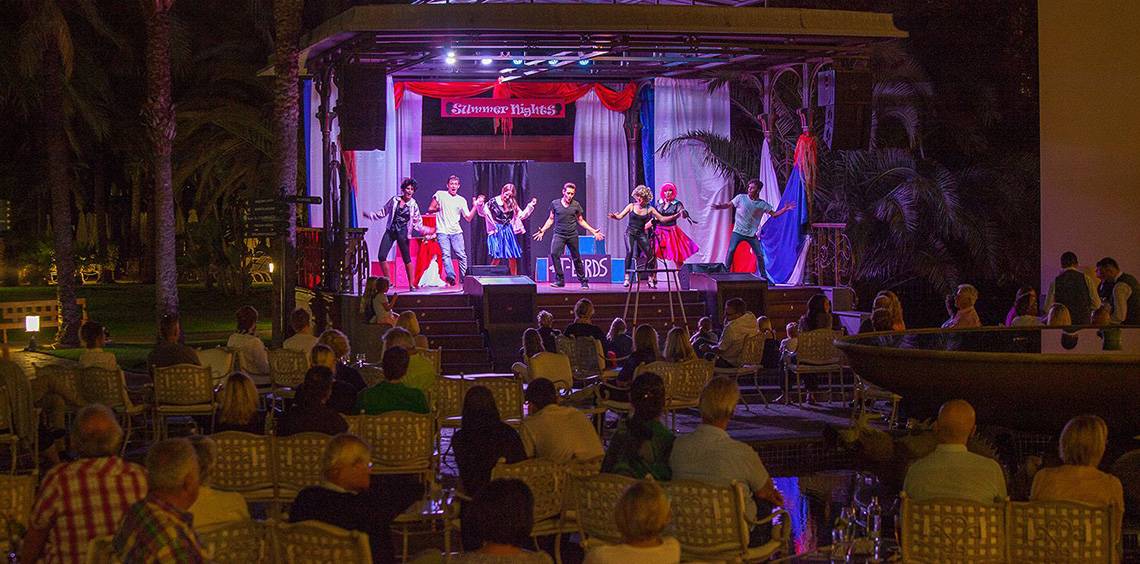 Eine Gruppe von Künstlern führt eine Show auf der Bühne des Lopesan Costa Meloneras Resorts & Spa auf, während das Publikum zuschaut.