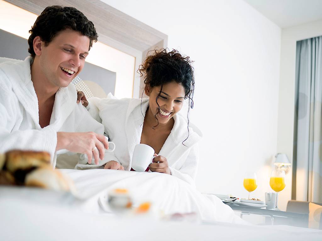 Ein Paar genießt Frühstück im Bett im Novotel London Blackfriars, beide tragen weiße Bademäntel, mit Kaffee, Orangensaft und Frühstücksbuffet im Hintergrund.