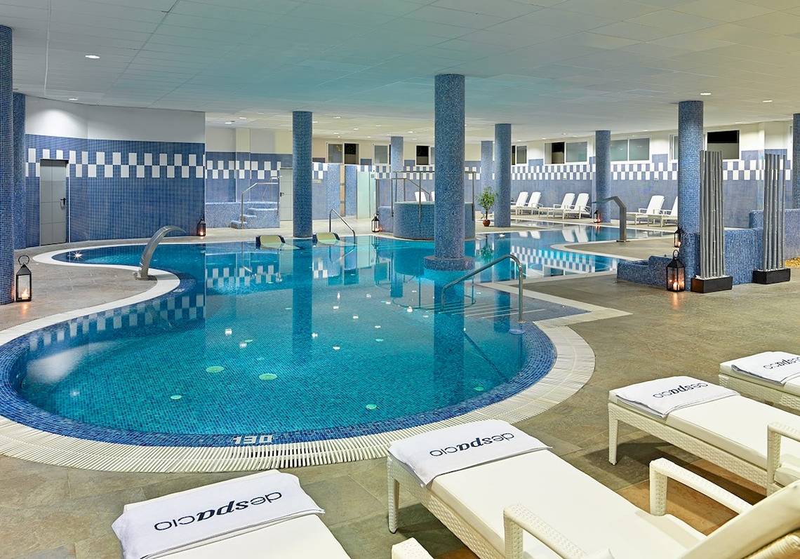 Indoorpool des H10 Estepona Palace mit Loungebereich und weißen Liegen.