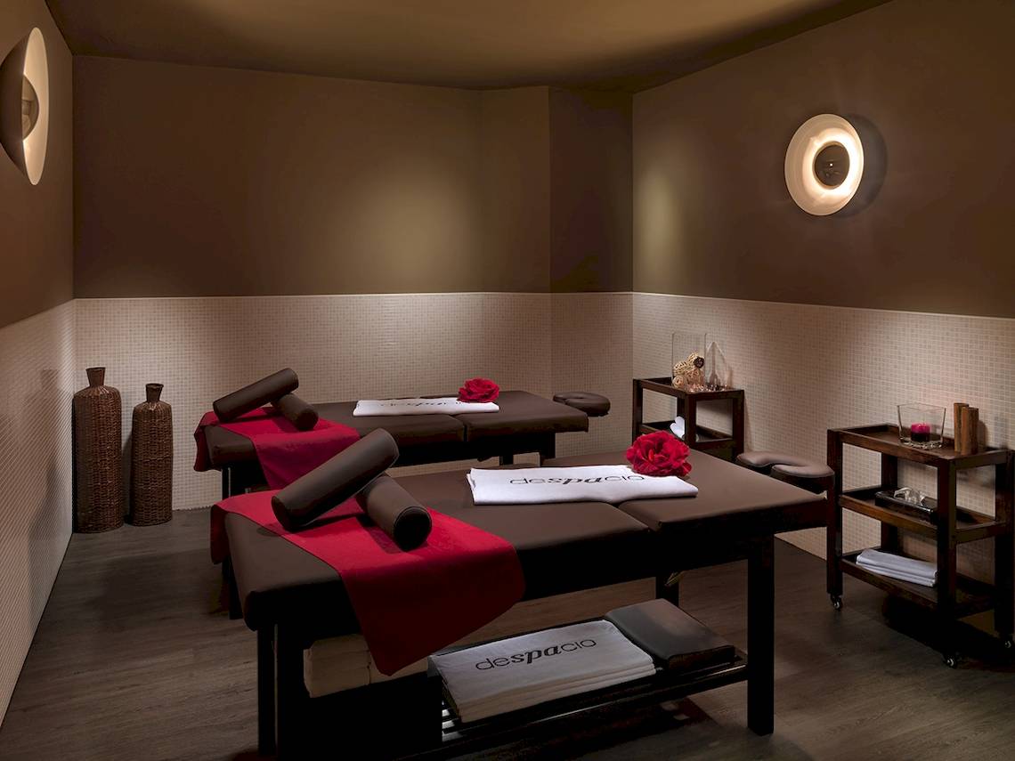 Spa-Behandlungsraum im H10 Estepona Palace Hotel mit zwei Massagetischen, roten Decken, und zwei Rosen.