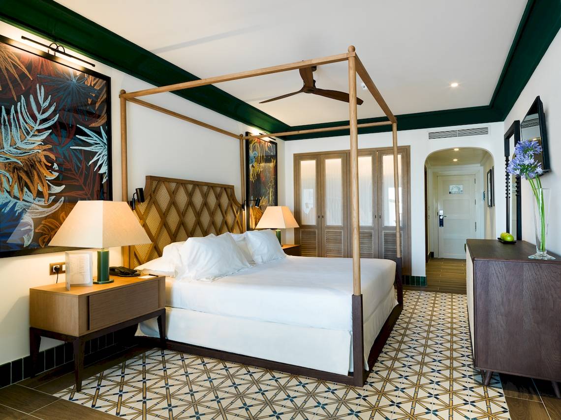 Schlafzimmer im H10 Estepona Palace mit Himmelbett, weißen Laken, zwei Nachttischen und tropischen Wandgemälden.
