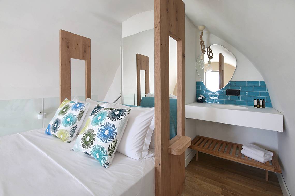 Innenansicht eines Hotelzimmers im Domes Noruz Chania, Autograph Collection, mit einem Bett, blauen Kissen und einem Badezimmerspiegel.