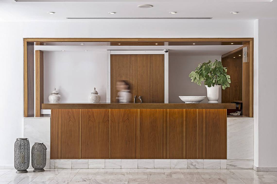 Empfangsbereich des Grecian Sands Hotels mit modernem Holzschalter, Marmorboden und Pflanzendekor.