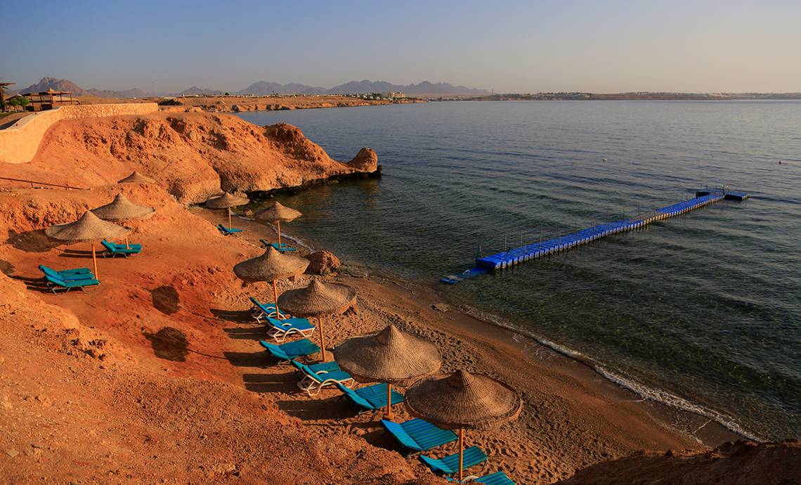 Strand des Sharm Club Beach Resorts mit Sonnenliegen, Sonnenschirmen und einer Brücke im Wasser.