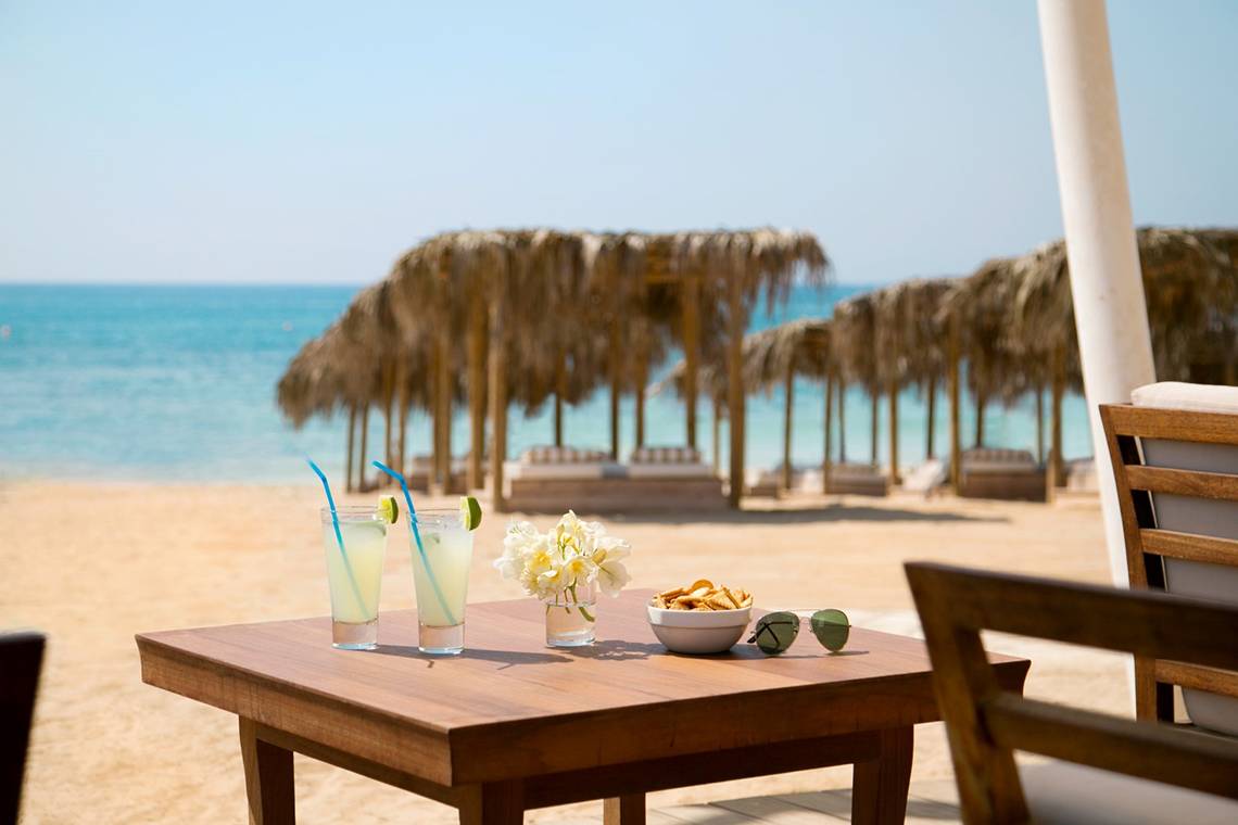 Sonnenterrasse des Asterias Beach Hotels mit Getränken und Blumen auf einem Tisch am Strand.