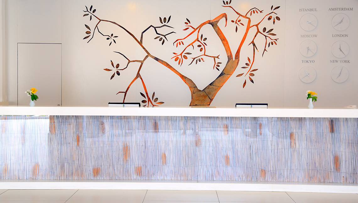 Rezeptionsbereich des Labranda TMT Bodrum mit einer Wandgestaltung, die einen Baum darstellt, und zwei Uhren, die verschiedene Städte darstellen.