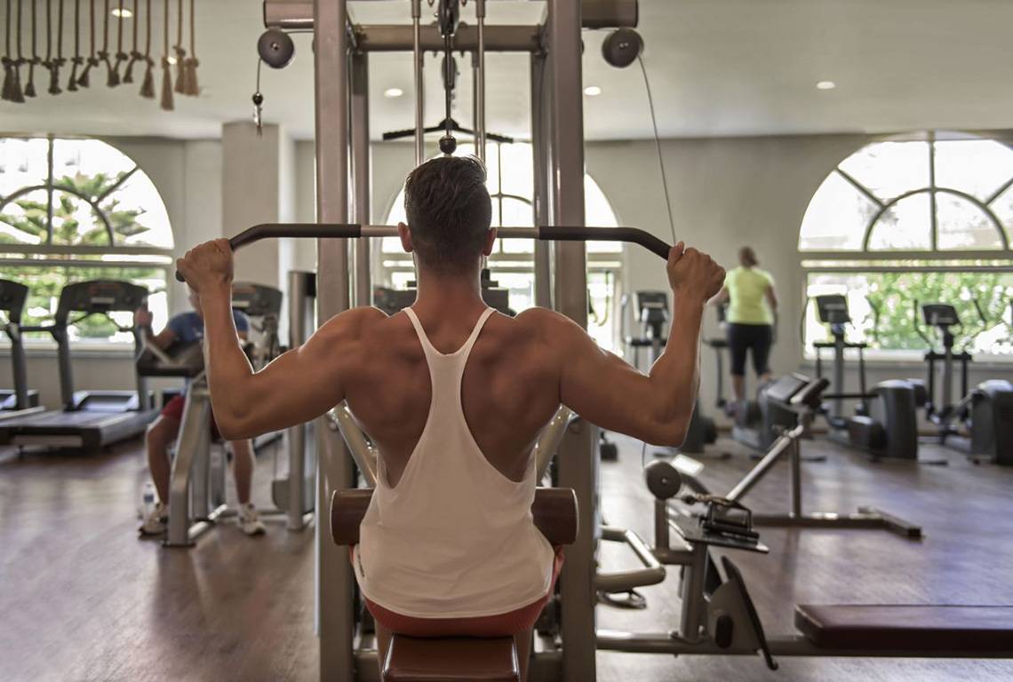 Ein Mann arbeitet im Fitnessstudio an einem Gerät, während zwei weitere Personen im Hintergrund trainieren.