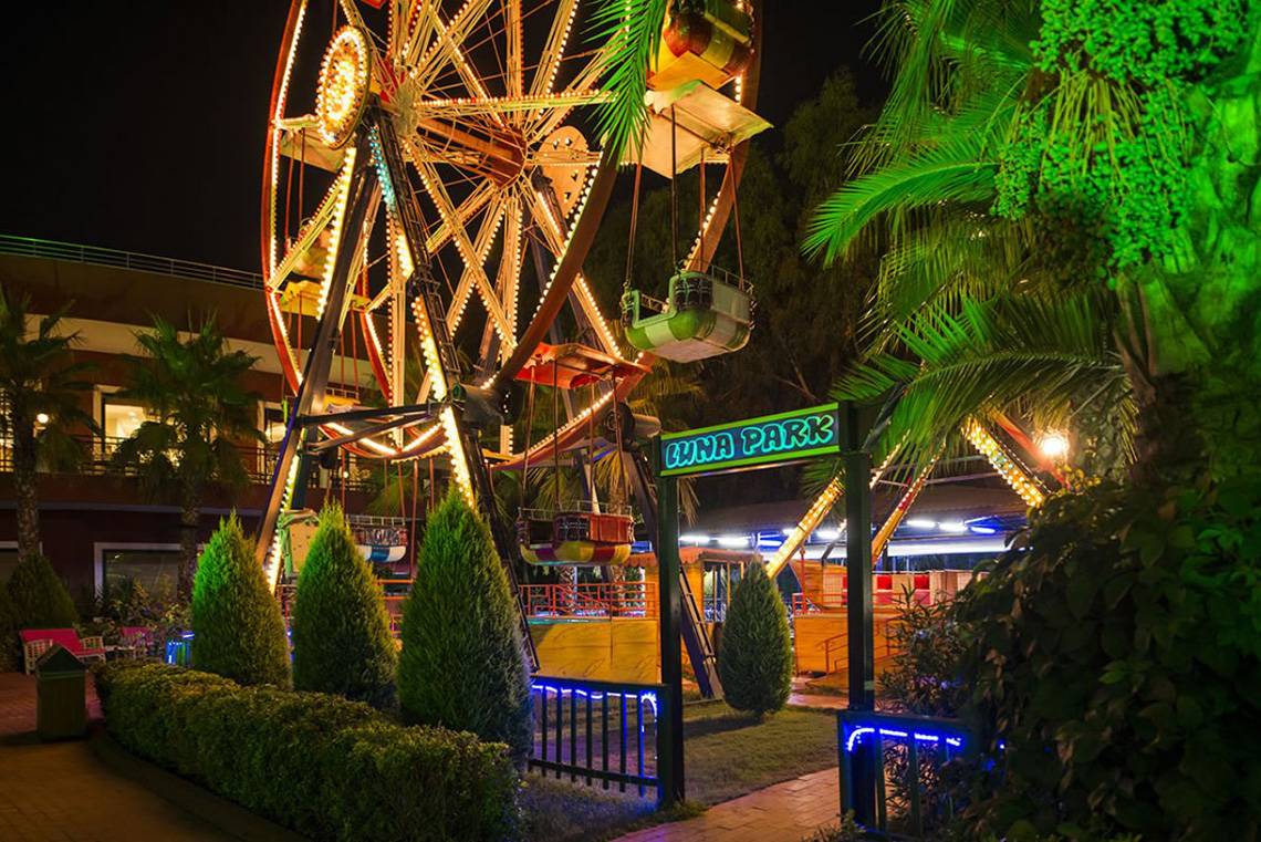 Beleuchtetes Luna Park im Delphin Palace Hotel bei Nacht mit Fahrgeschäften und Palmen.