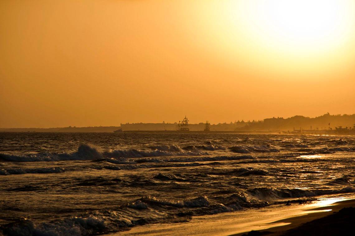 Sonnenuntergang am Strand mit Wellen und im Hintergrund eine Stadtsilhouette
