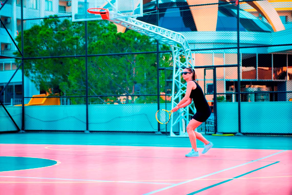 Eine Frau spielt Tennis auf einem Sportplatz, der mit einem Basketballkorb im Hintergrund gestaltet ist.