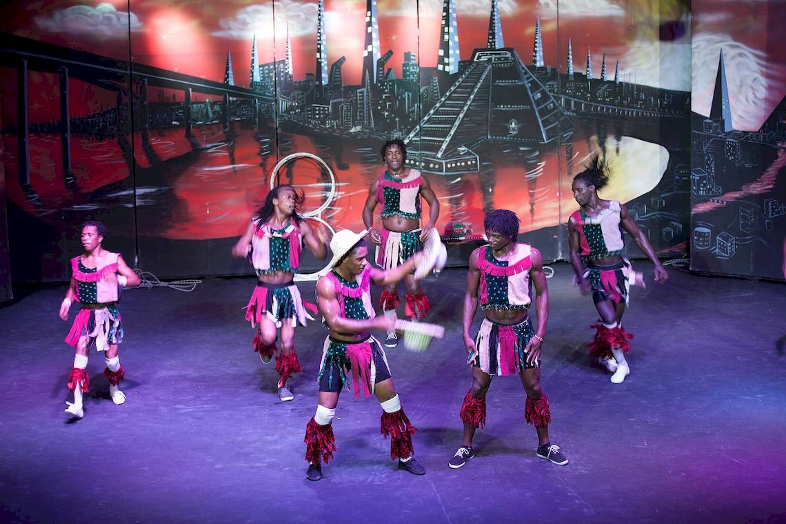 Afrikanische Tanzgruppe auf einer Bühne mit farbenfroher Kulisse und rotem Hintergrund.
