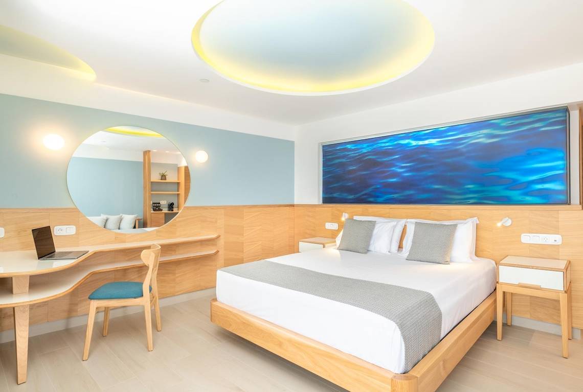 Innenansicht eines Zimmers im Lyttos Mare Hotel mit Bett, Schreibtisch, Stuhl und Aquariumswand.