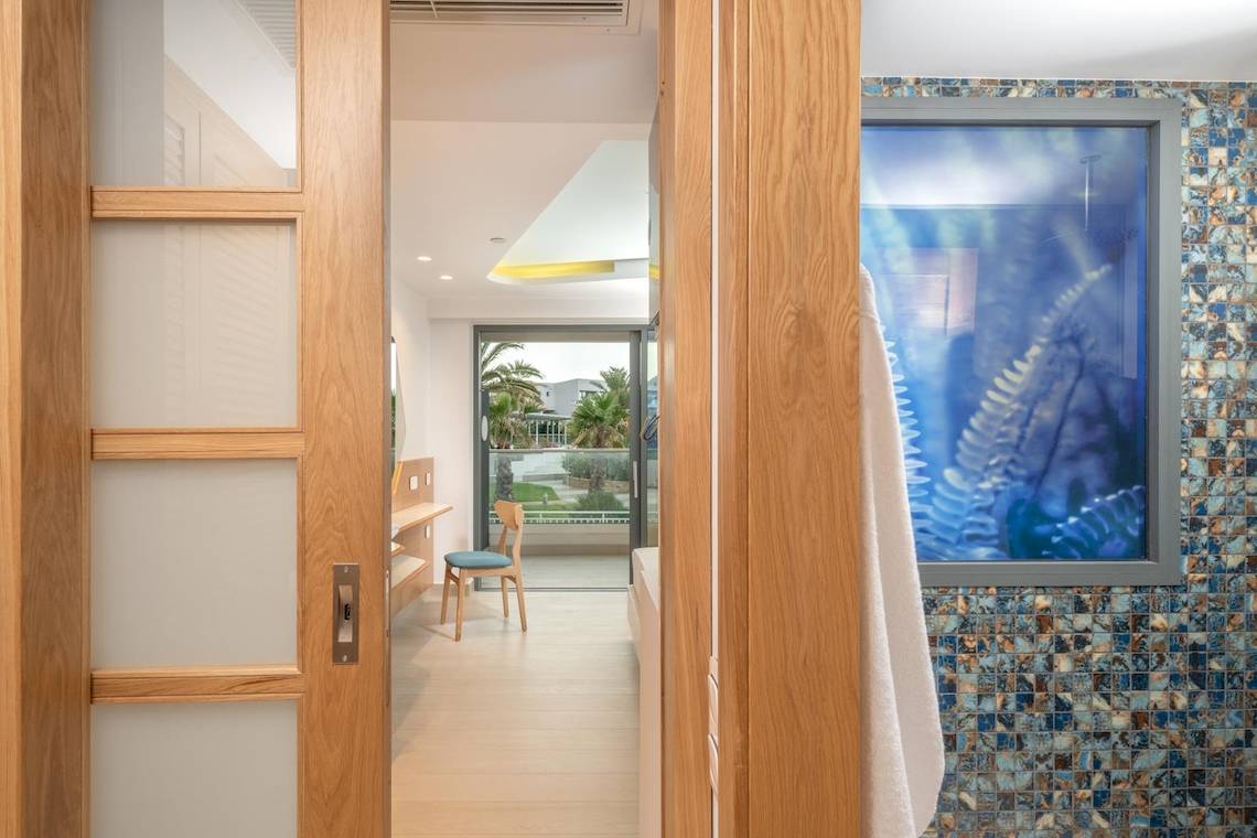 Innenansicht eines Zimmers im Hotel Lyttos Mare mit offenem Holztür, Holzmöbel und Ausblick auf Garten