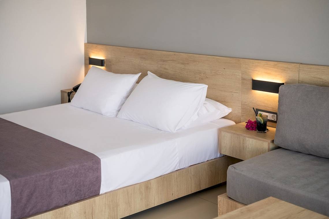 Schlafzimmer im CHC Galini Palace Hotel mit Doppelbett, weißen Kissen, Nachttisch und Beleuchtung.
