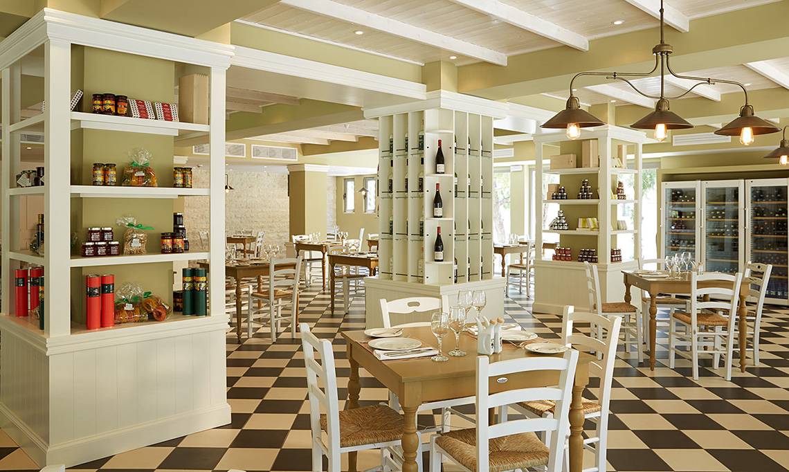 Innenansicht des MarBella Corfu Restaurants mit schachbrettartigem Boden, weißen Möbeln und Regalen mit Produkten.
