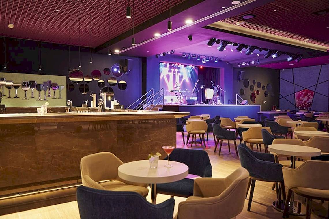 Innenansicht der Bar im Riu Dubai Hotel mit Tisch, Stühlen und einer Bühne.