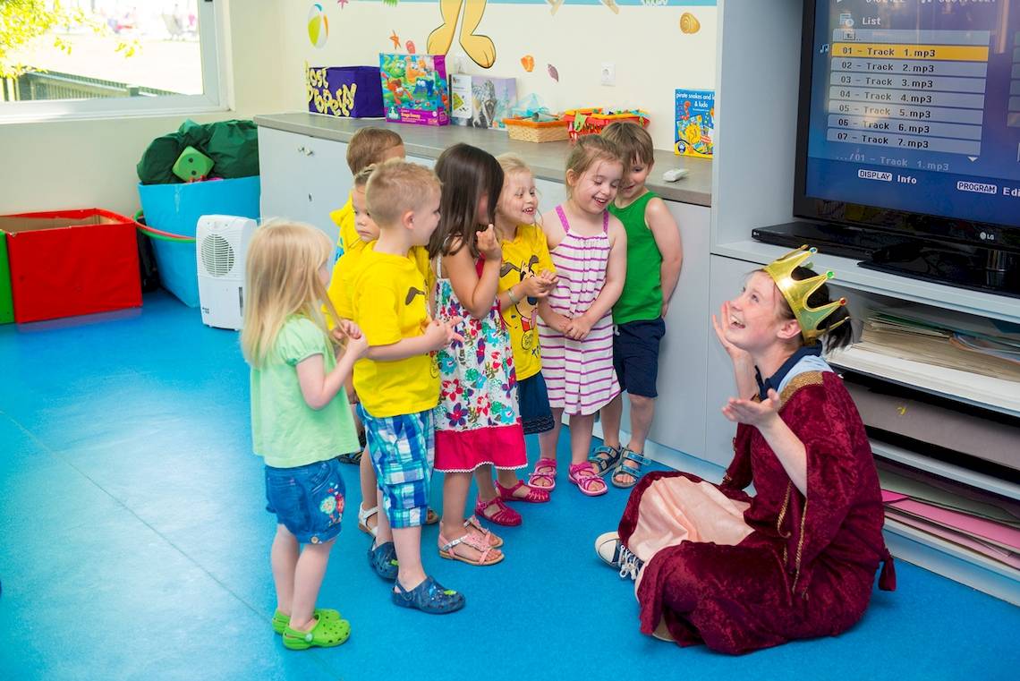 Gruppe von Kindern in einem Spielraum, eine Frau in einer Krone sitzt und singt vor ihnen.