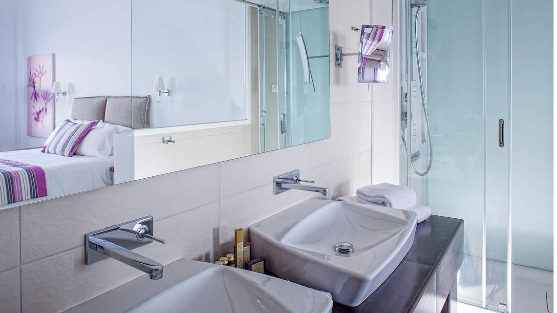 Badezimmer mit einem großen Spiegel und zwei Waschbecken, reflektiert ein Bett und eine Dusche.