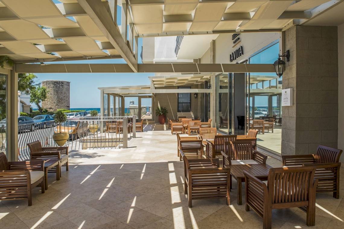 Terrasse des Mitsis La Vita Beach Hotels mit Holzmöbeln und Blick auf das Meer