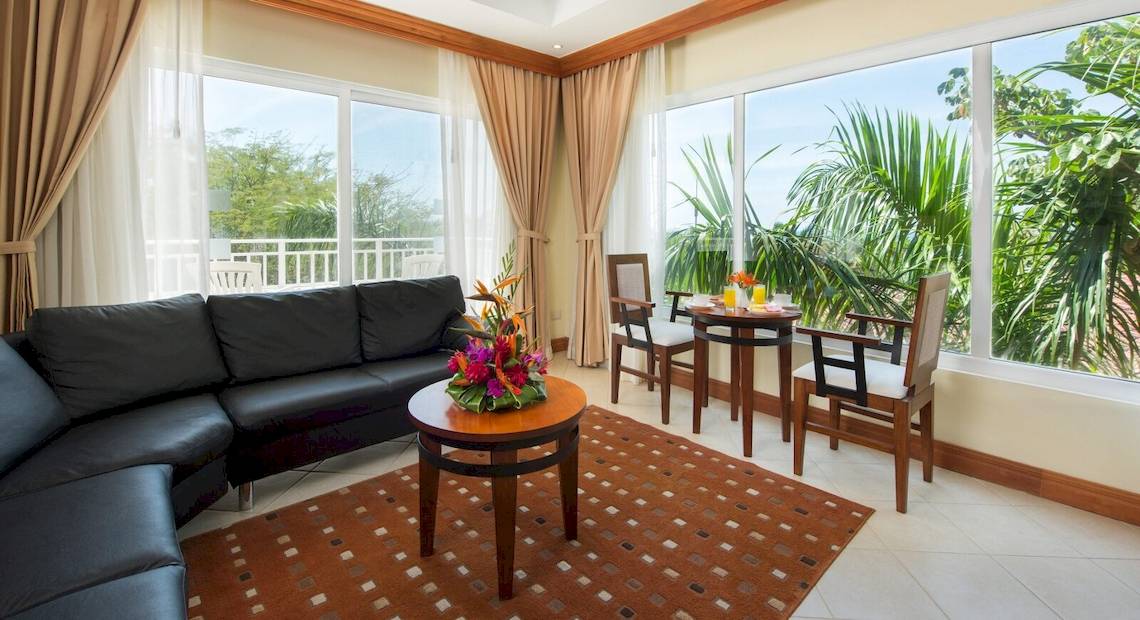 Wohnzimmer im Occidental Tamarindo Hotel mit schwarzem Sofa, Tisch und großem Fenster mit Blick auf Palmen