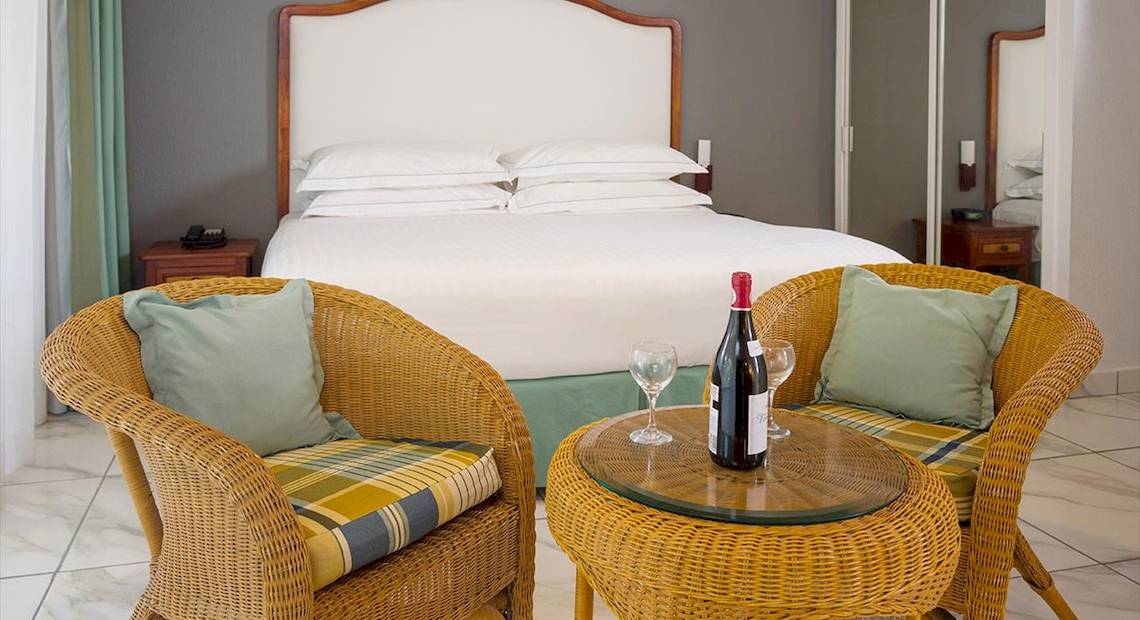 Ein elegantes Zimmer im Occidental Tamarindo Hotel mit zwei Rattanstühlen, einem Tisch, einer Weinflasche und zwei Weingläsern neben einem großen Bett.