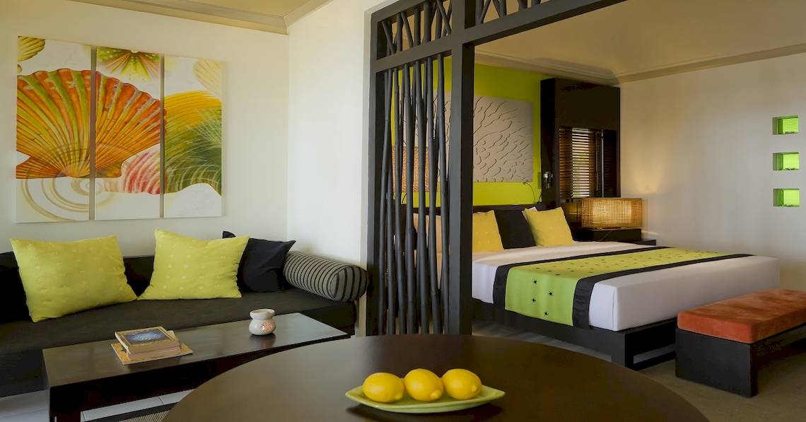 Innenansicht eines Zimmers im Dhawa Ihuru Hotel mit Bett, Couch, Tisch und Gemälden an der Wand.
