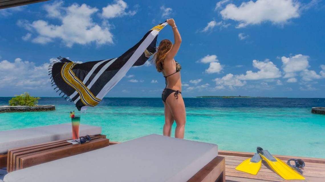 Eine Frau in Bikini am Strand des Lily Beach Resort & Spa, das Handtuch haltend, mit Blick auf das Meer.