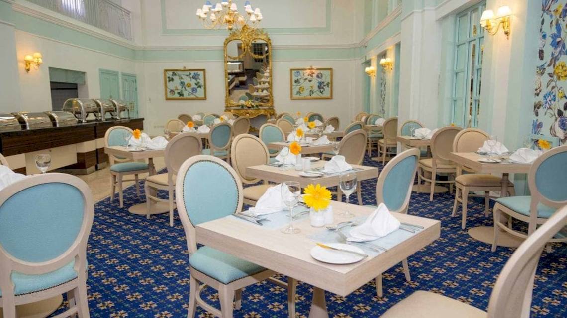 Speisesaal des AX The Victoria Hotels mit blauen Teppich, weißen und blauen Stühlen, Tischdecken und Blumengestecken.