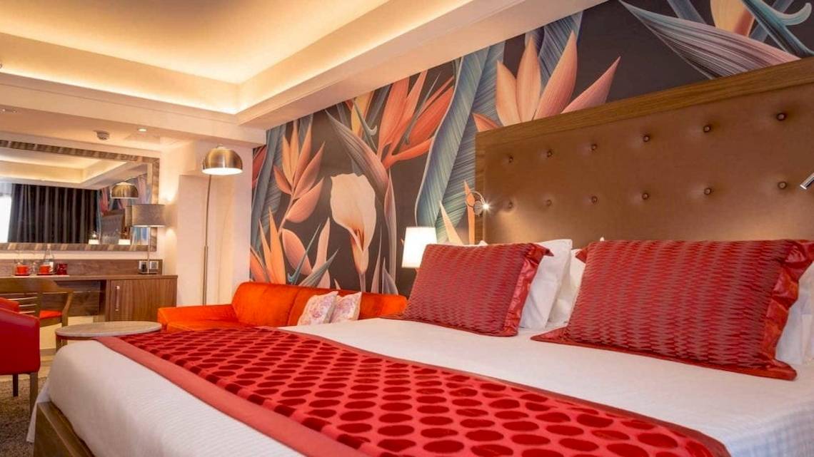 Schlafzimmer im AX The Victoria Hotel mit tropischen Wandmalereien, einem großen Bett, zwei roten Stühlen und einem Couchtisch.