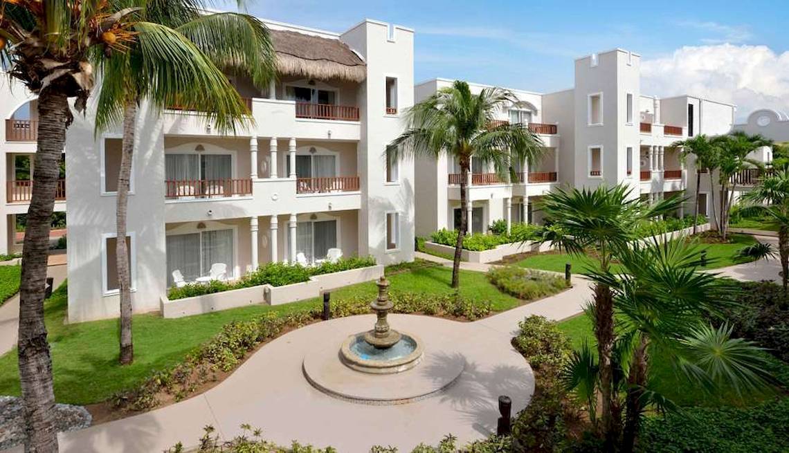 Außenansicht des weißen Catalonia Playa Maroma Hotels mit Palmen und einem Brunnen