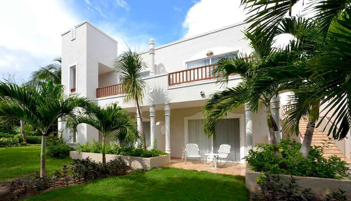 Außenansicht des weißen Hotels Catalonia Playa Maroma mit Balkon, Terrasse und Palmen.