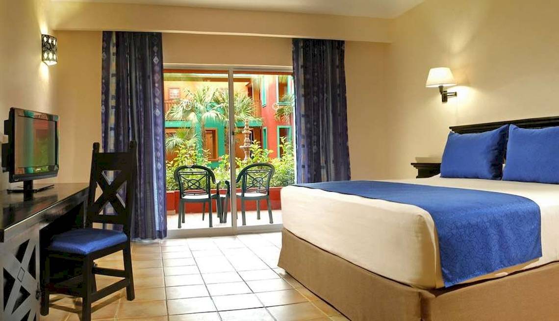 Ein Hotelzimmer mit Balkon, Fernseher, Schreibtisch und zwei Stühlen vor einem Glasfenster.