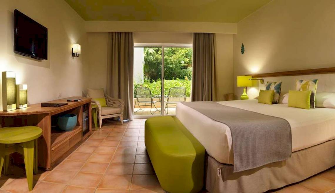 Hotelzimmer im Catalonia Playa Maroma mit Doppelbett, grünen Stühlen und Tisch, Fernseher und Balkonzugang.