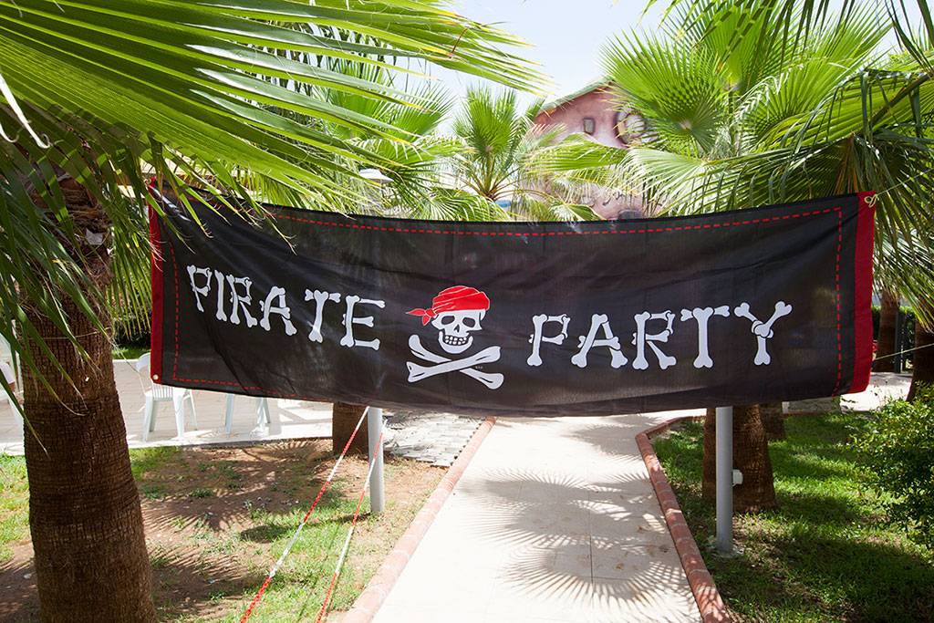 Schwarzes Banner mit dem Text 'Pirate Party' und einem Totenkopf, befestigt zwischen zwei Palmen.