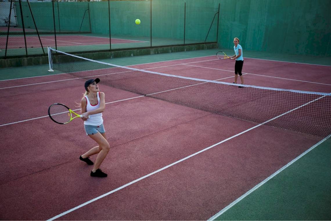 Zwei Frauen spielen Tennis auf einem roten Tennisplatz mit einer Netz und einem Tennisball in der Luft.