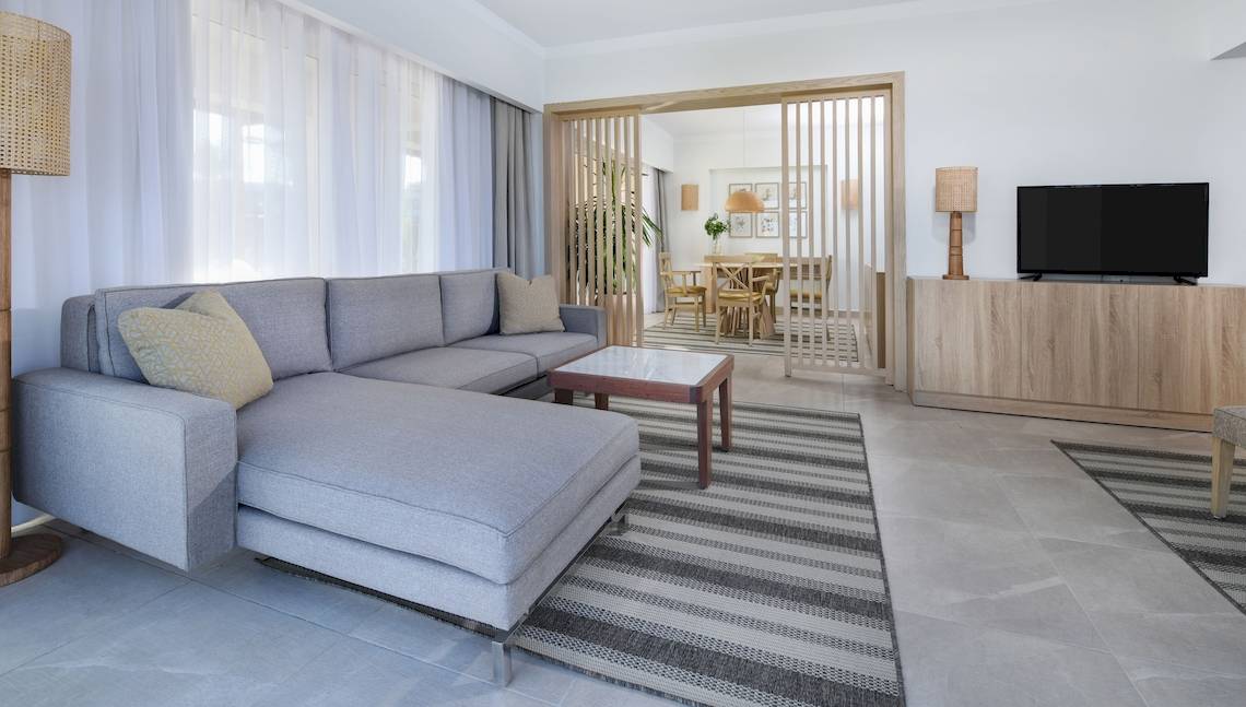 Wohnzimmer des Iberotel Costa Mares Hotels mit grauem Sofa, Couchtisch, Fernseher und Tisch mit Stühlen.
