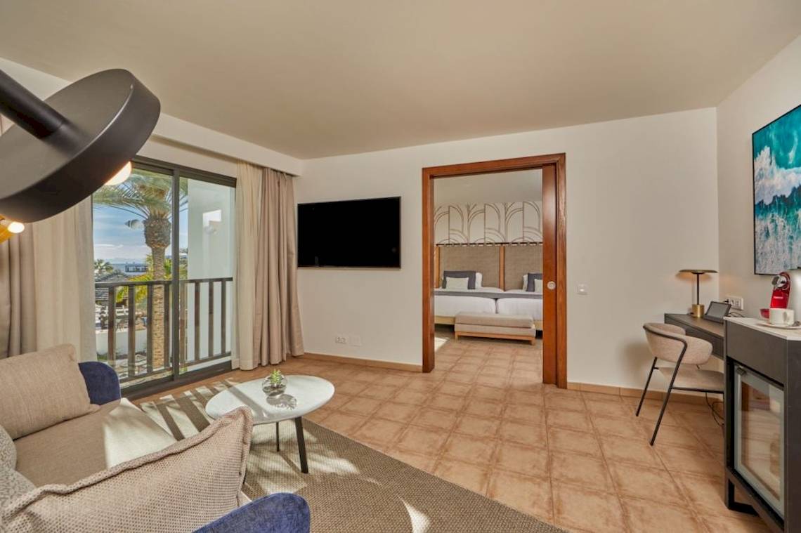 Innenansicht der Suite im Secrets Lanzarote Resort & Spa mit Wohnzimmer, Couch, Tisch und Balkon.