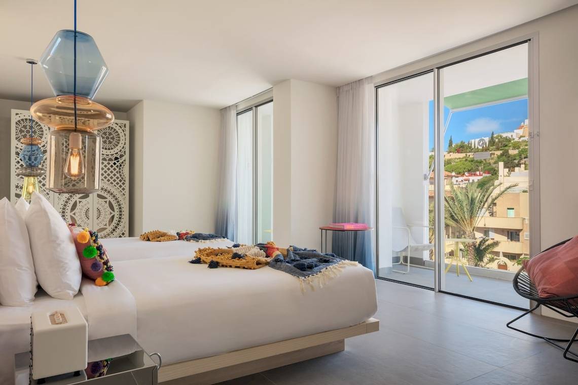Innenansicht eines Zimmers im W Ibiza Hotel mit zwei Betten, Decken, Kissen und einem Balkon.