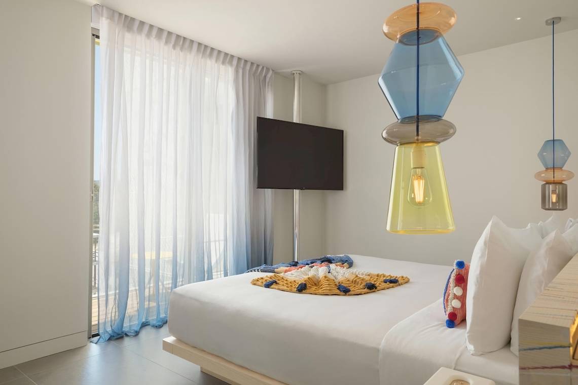 Ein modernes Hotelzimmer im W Ibiza mit einem großen Bett, farbigen Deckenleuchten und einem Fernseher.